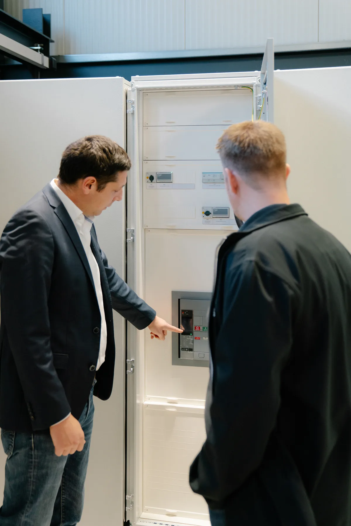 Zwei Männer vor einem geöffneten Schaltschrank, einer zeigt auf ein eingebautes Prüfgerät